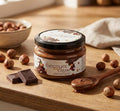 Jar of chocolate cream spread on a wooden surface with nuts and chocolate pieces.